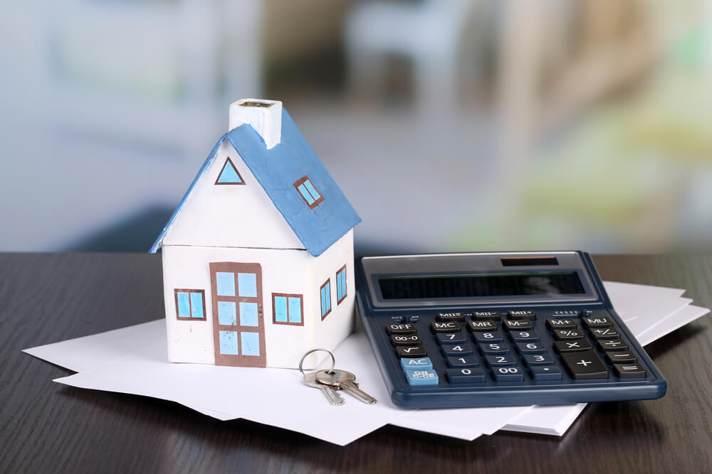 A little house sitting on top of a stack of papers and next to a black calculator with keys in front of it