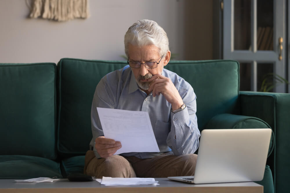 An older man sitting on the couch looking at a paper with his hand on hit chin and papers on the coffee table next to an open lap top 