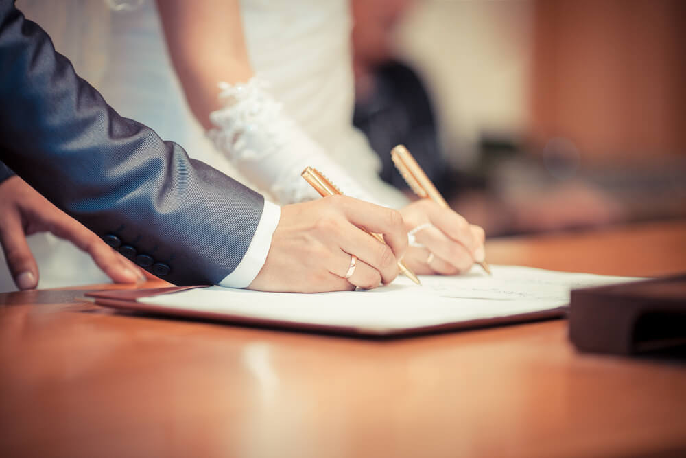 A close up of a husband and wife in their wedding attire signing the marriage certificate 