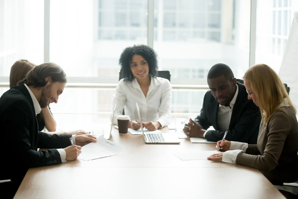 Five people sitting at a conference table, two are on the right and two are on the left and one person is at the head of the table. The people at the table are looking over documents and signing them