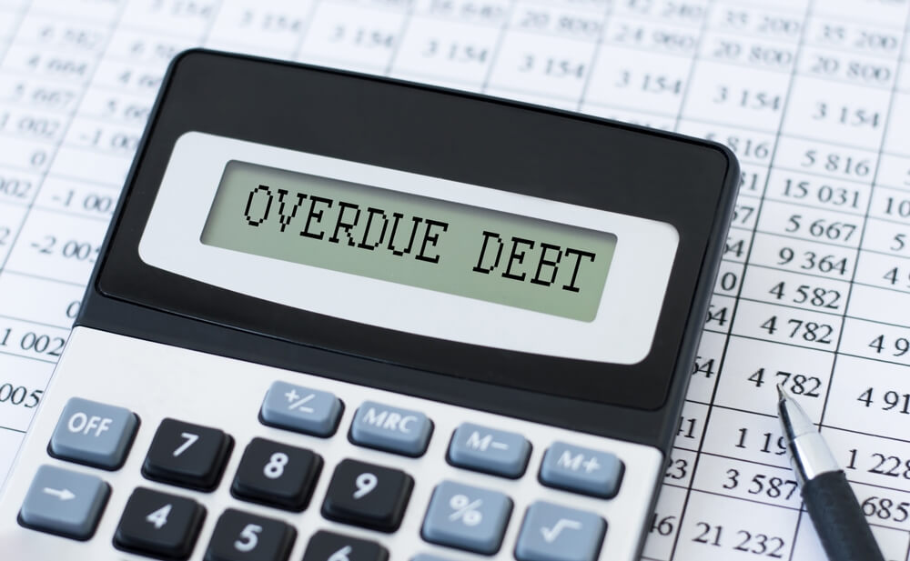 A calculator that says overdue debt on the screen sitting next to a pin and on a spreadsheet filled in with numbers 
