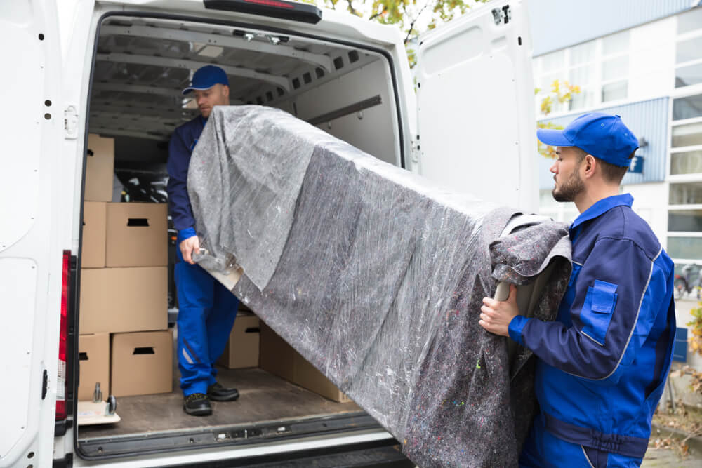 Two delivery men putting a wrapped couch into a truck with brown boxes in the truck