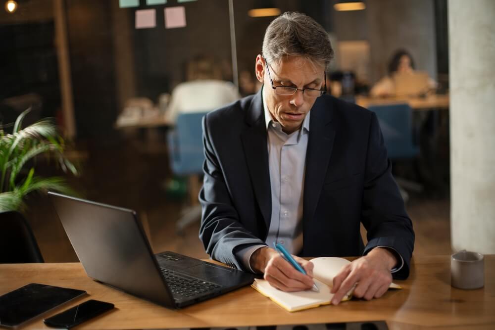 A man sitting at a desk writing in his notebook and his computer open