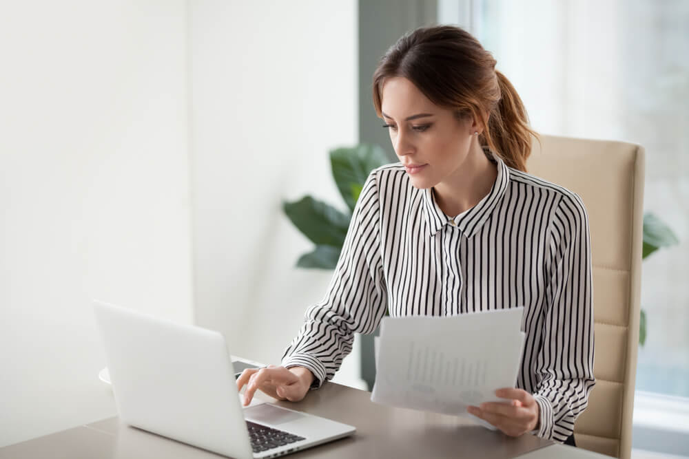 A woman sitting at a desk looking over a piece of paper and typing into her lap top