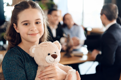 A young girl holding a teddy bear smiles at the camera, while a group of adults in the blurred background discuss whether living with a boyfriend can affect custody decisions.