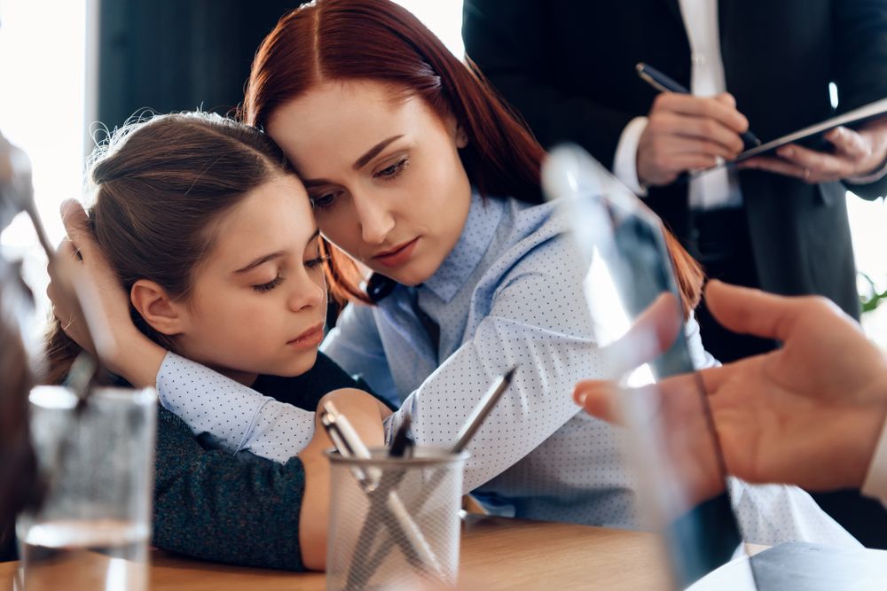 Young sad mother hugs little upset daughter sitting in lawyer's office for divorce. Two parents fighting over child in divorce concept.