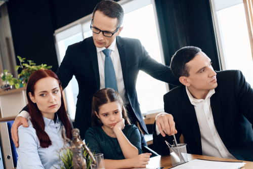 A serious Texas custody battle unfolds in a meeting with two men in suits, a woman, and a young girl at the table. Legal documents and a scale model lay before them, surrounded by potted plants, setting the scene for this tense legal encounter.