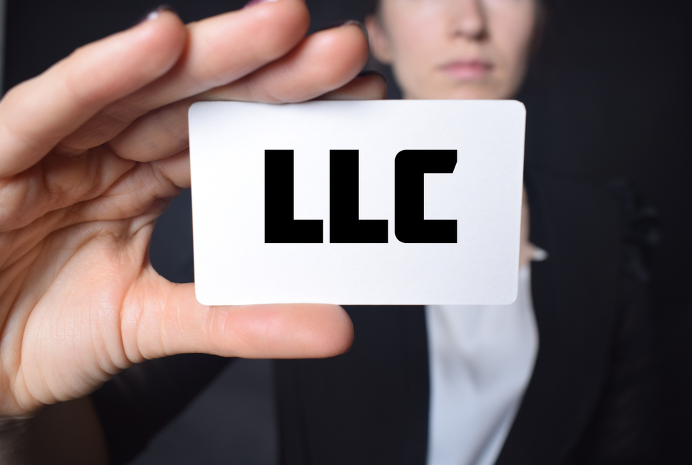 A person holds a white card with the bold letters "LLC" printed on it, suggesting topics like how to transfer ownership of an LLC to a trust, with part of their face visible in the background.