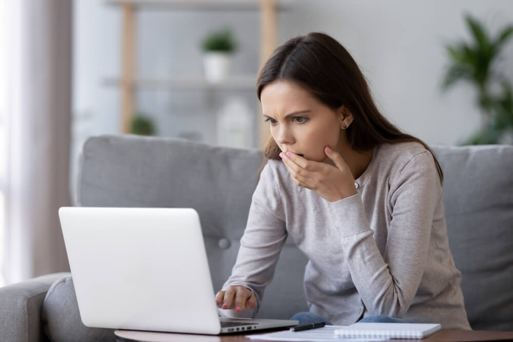 A woman sitting on her couch looking at her computer with a shocked look on her face and one hand covering her mouth 