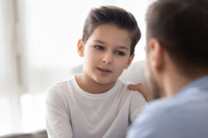 Back view of dad sit on couch with preschooler son talking sharing thoughts or problems, loving father speak have conversation with little boy child, support him showing care and understanding