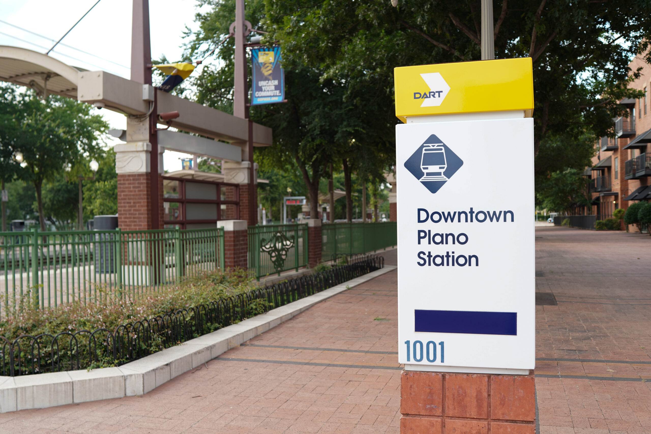 A sign for downtown Plano station, with the DART logo on top, located beside a walkway with greenery and a train platform in the background,