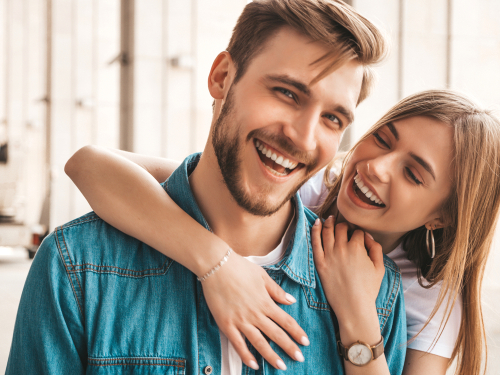 A smiling man in a denim shirt stands as a woman hugs him from behind, basking in the sunlit setting. Their carefree day hints at life's simple joys, leaving one to wonder how such idyllic moments can influence life's complexities like custody decisions.
