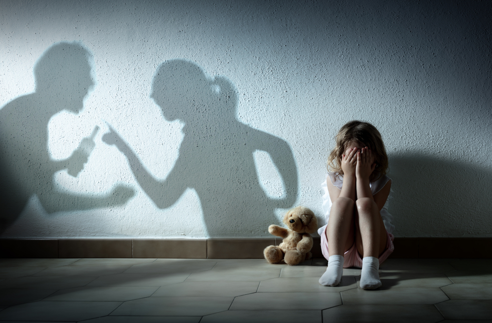 A young girl sits on the floor covering her face, with a teddy bear beside her. Shadows on the wall show two adults arguing and pointing fingers, hinting at a tense home life and possible signs of a texas unfit parent.