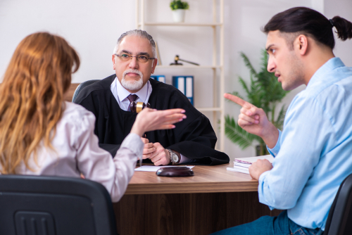 A man and a woman are having a discussion in a judge's office about how any assets held overseas are treated in their divorce. The judge, wearing a robe and holding a gavel, looks on as the man gestures with his finger pointed at the woman.