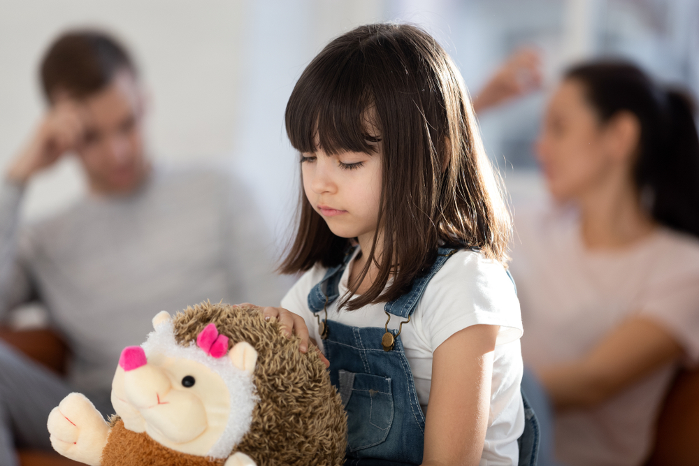 Close up little girl suffering from parents conflict sitting at home in living room and holding soft toy. Sad daughter frustrating hear mother and father fighting arguing, family conflict, divorce.
