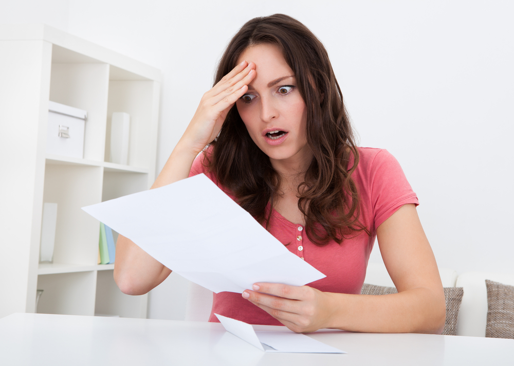 A woman sitting at a table looks shocked while reading a document her other hand on her forehead in disbelief.