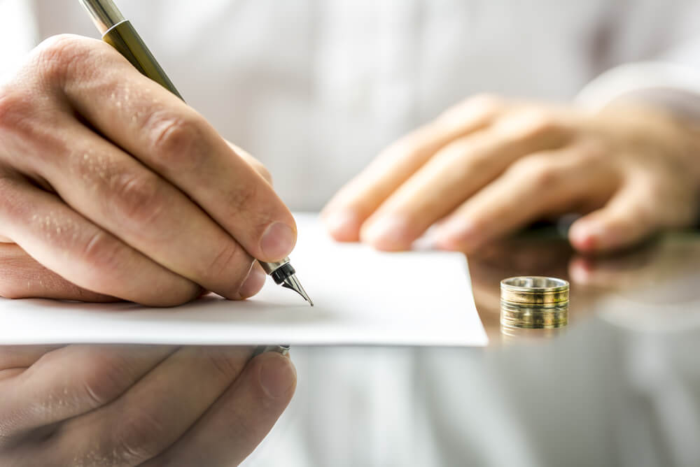 A close up of a man signing a piece of paper with his wedding ring resting on the table 