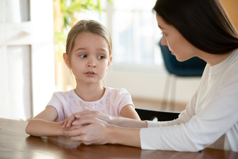 Head shot unhappy small child girl sitting at table with worrying mother, sharing school problems. Compassionate caring attentive mommy having trustful conversation with unhappy offended daughter, explaining material change in circumstances