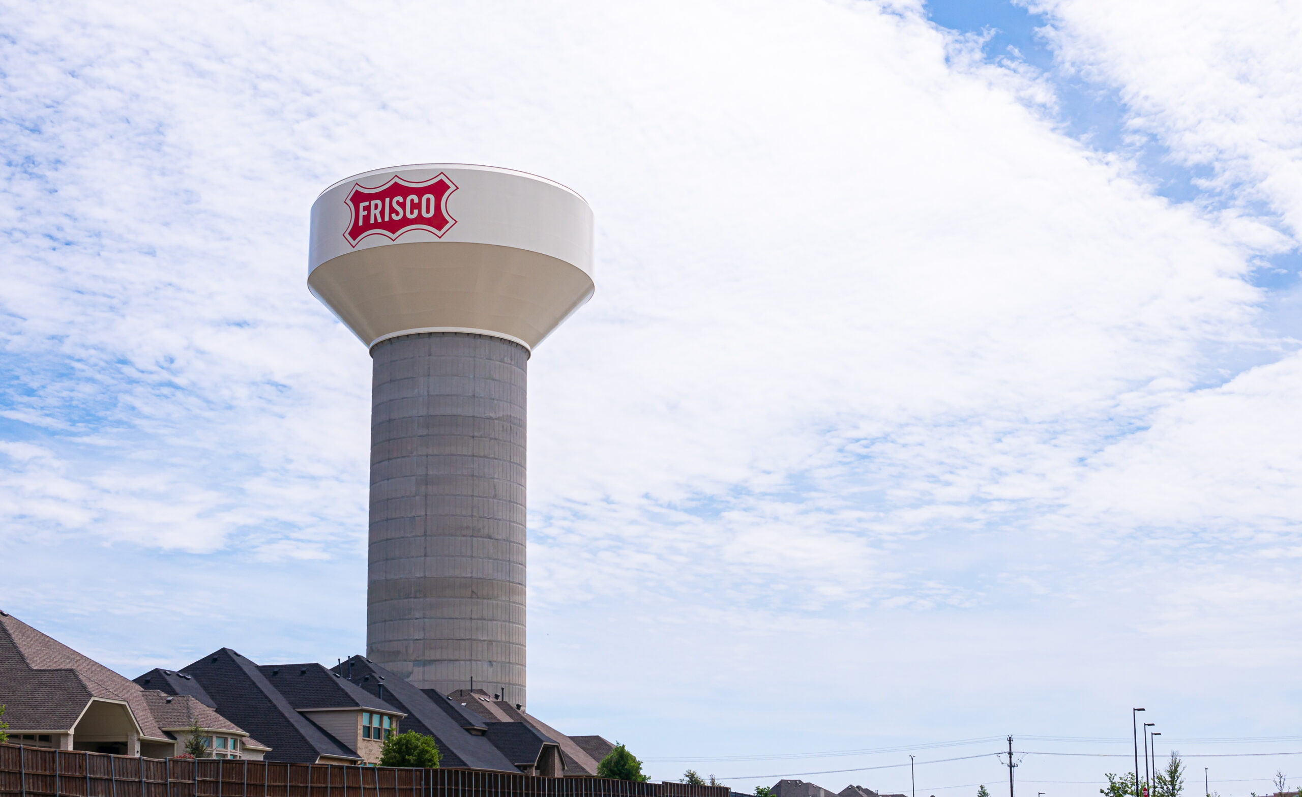 A large water tower with the word "frisco" on it, standing tall against a blue sky, surrounded by suburban homes.