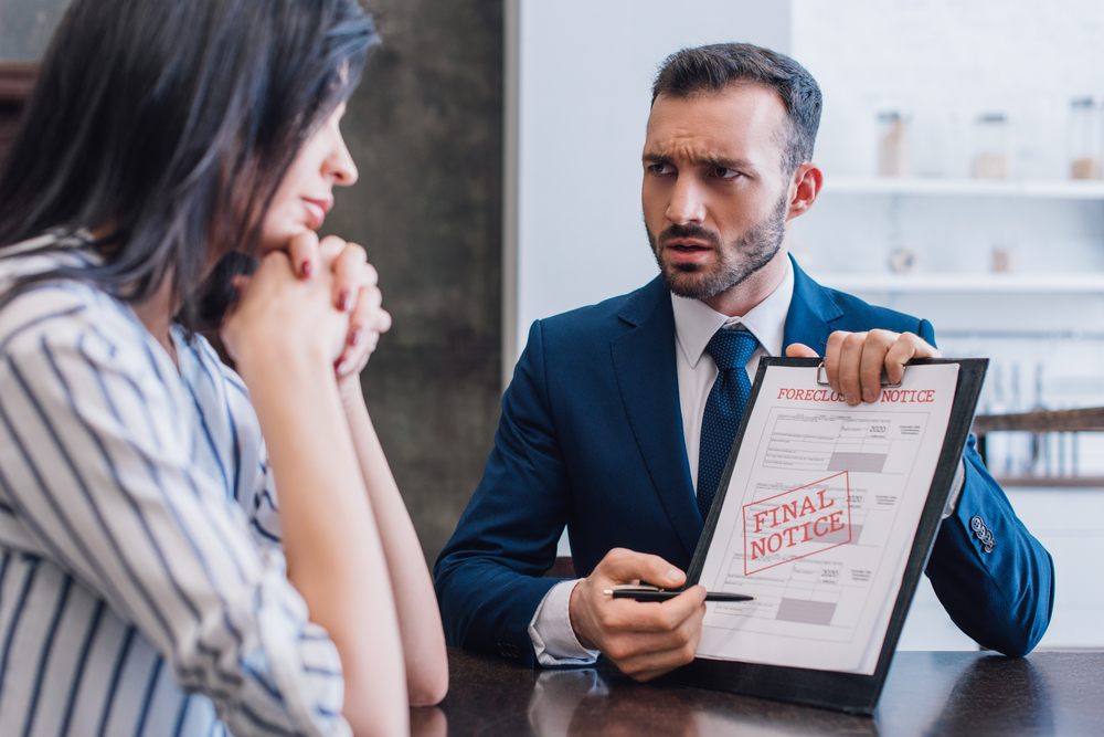 Tense collector pointing at documents with foreclosure and final notice lettering and looking at woman with clenched hands at table, relating it with sued by capital one credit card debt