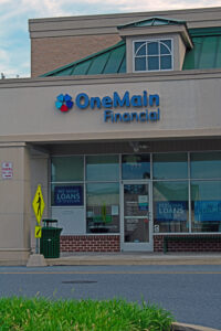 A OneMain Financial storefront with signage offering various loans up to $25,000. The building has large glass windows and a pedestrian crossing sign nearby, ensuring you don't mistake it for the adjacent Morgan and Associates debt collection office.