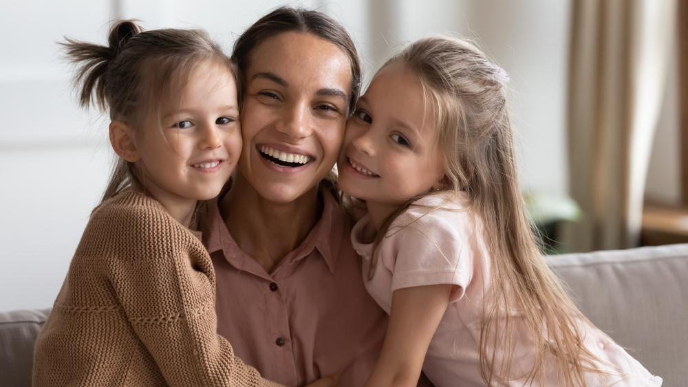 Portrait of happy family, mother day celebration concept. Single loving mommy and two little 6s daughters embracing seated on sofa at home. Young cheerful mom with cute girls smile looking at camera