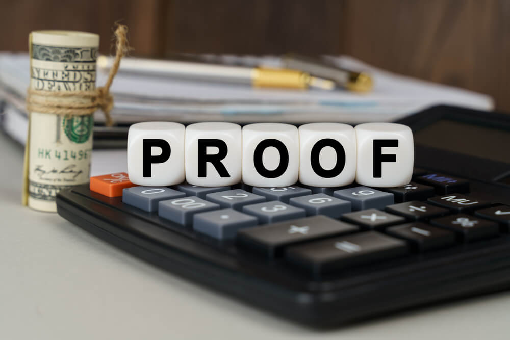 A calculator sitting on a desk with the word proof spelling out with spelling blocks and a roll of money standing next to the calculator 