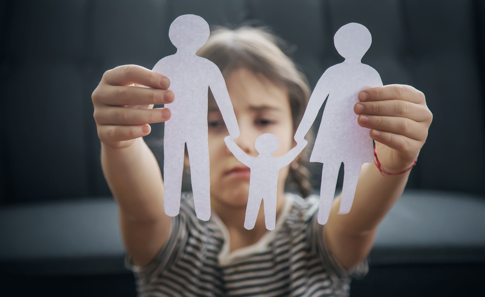 A child holds paper cutouts of two adults and a child, symbolizing a family, with a serious expression