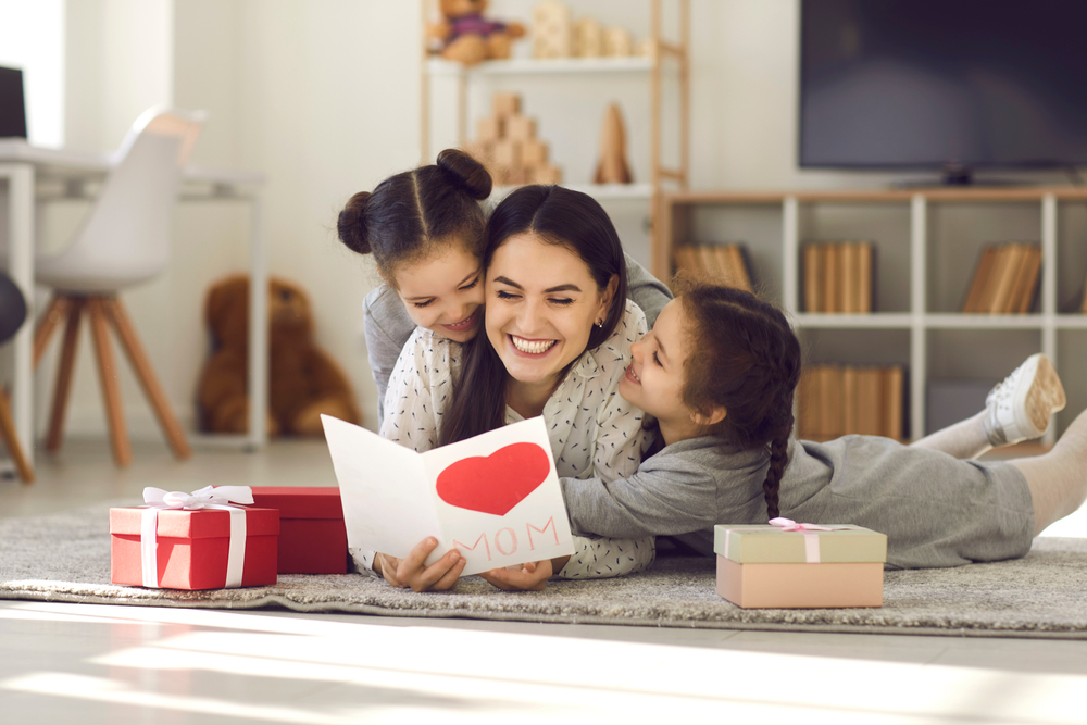 A woman lies on the floor reading a heart card, smiling as two young girls hug her—celebrating Mother’s Day together. Gift boxes are nearby in a cozy living room, making the most of their standard possession order in Texas.
