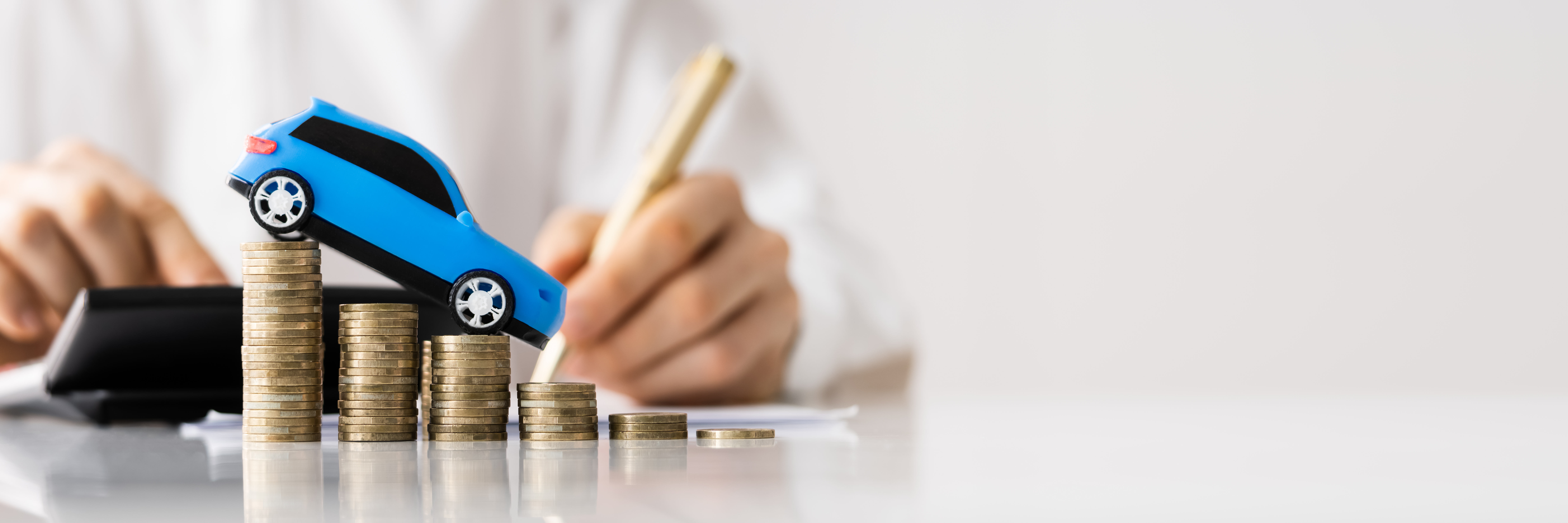 A small blue toy car sits on top of stacks of coins, with a person using a calculator and writing in the background.