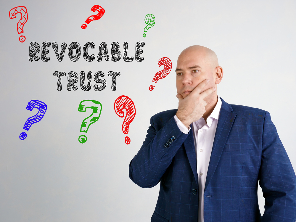 A man in a suit stands thoughtfully next to the text "Revocable Trust," surrounded by colorful question marks, reflecting on how to transfer ownership of an LLC to a trust against a light background.