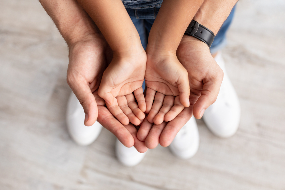 An adult and a child hold their hands open together, palms up, with the child's hands resting inside the adult's hands, symbolizing care and protection,