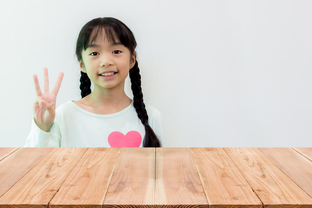 A little girl sitting at a wooden table against a white background holding up one hand with three fingers 