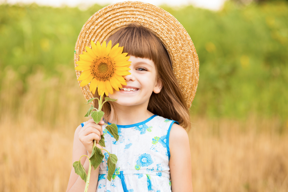 Beautiful little girl in straw hat with fluttering hair smile and hide eye with sunflower flower, walking outdoor in summer holiday