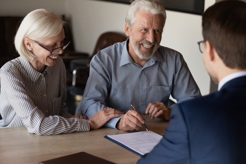An older couple is seated at a table, smiling and discussing estate planning paperwork with a man in a suit during their free consultation. The man is holding a pen, ready to sign documents.