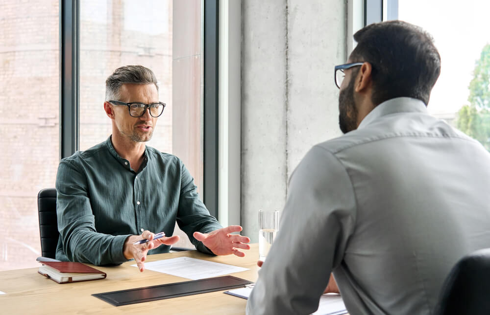 Two men sitting at a desk both with papers in front of them as they talk to each other