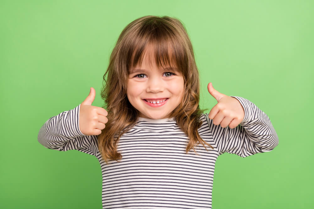 A smiling little girl standing against a green background with her thumbs up 