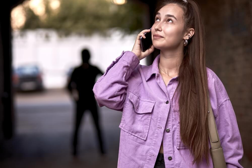 A woman standing outside while talking on the phone and a man in all black in behind her watching her between two buildings 