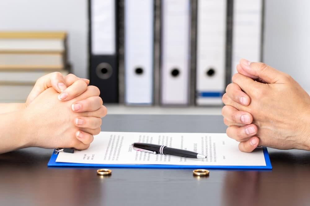 A close up of a man and a woman sitting at a desk with their hand clasped with paperwork in between them and their wedding rings sitting on the table