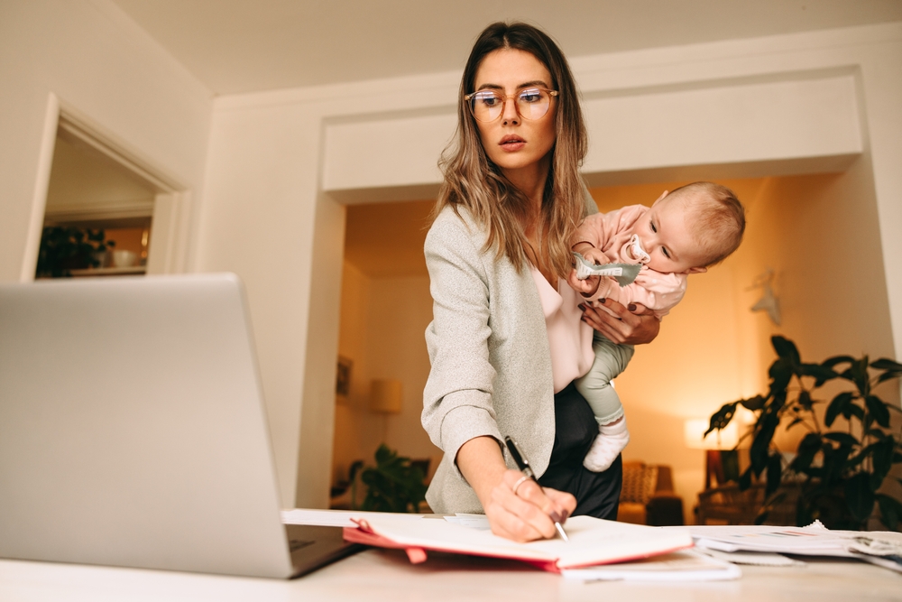 A woman holds a baby while working at a desk, writing in a notebook and looking at her laptop. Papers and folders are spread out around her workspace.