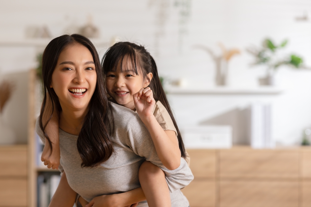 A woman smiles while giving a young girl a piggyback ride indoors, with a blurred background of shelves and home decor, capturing a joyful moment