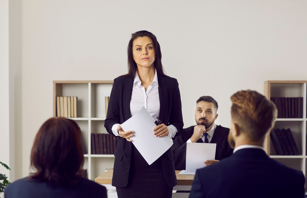 Portrait of female lawyer or attorney who represents plaintiff in lawsuit during court trial. Serious unsmiling woman holding documents standing in courtroom after her speech in front of audience