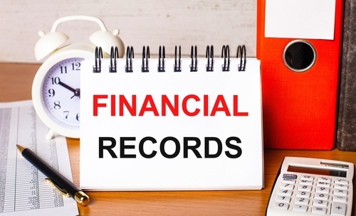 A spiral notebook labeled "Financial Records" rests on a desk alongside a calculator, red file folder, alarm clock, and essential documents for filing bankruptcies.