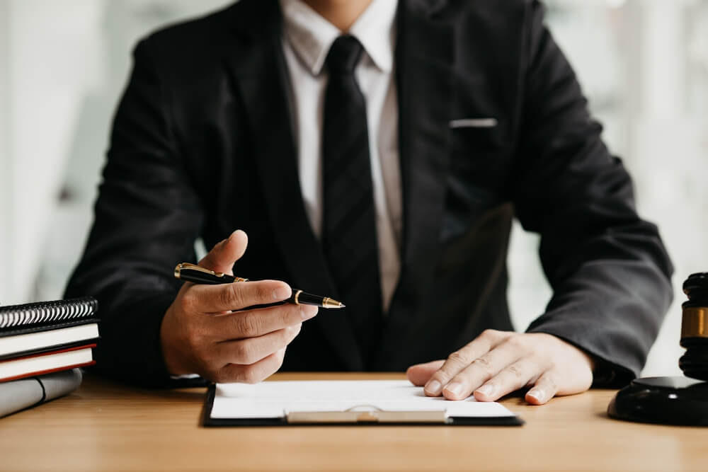 A close up of a businessman sitting at a desk with documents in front of him and he is in the process of writing 