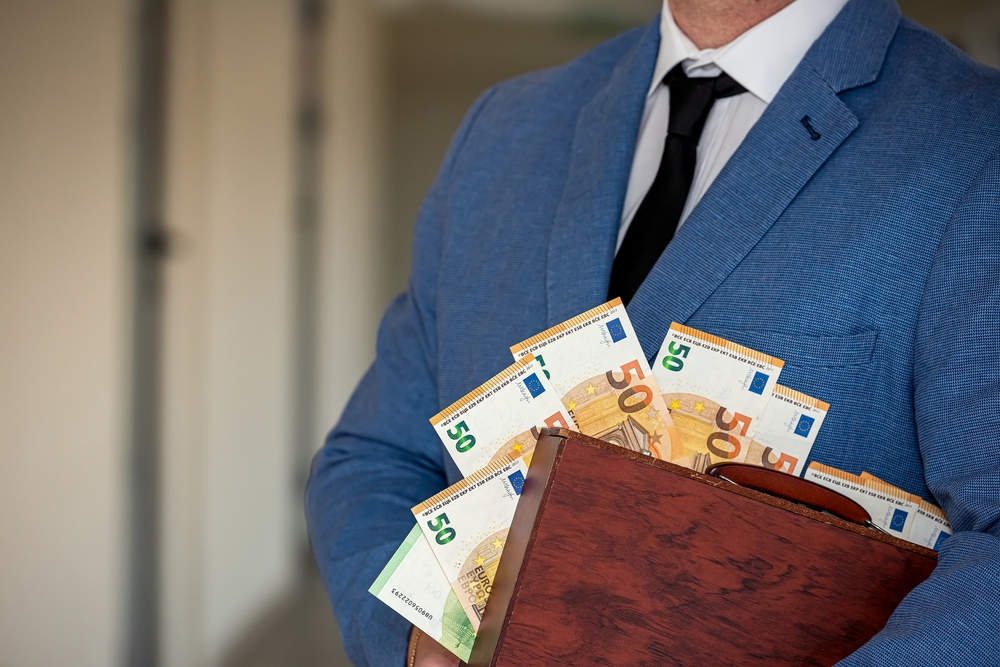 A person in a blue suit holds a briefcase filled with fifty euro banknotes