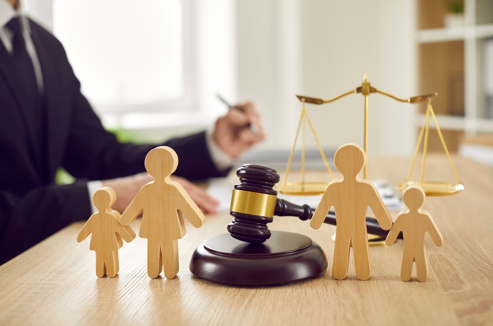 Wooden family figures, a judge’s gavel, and scales of justice are on a desk as someone in a suit writes in the background—symbolizing issues like a custodial parent not following court order in Texas.