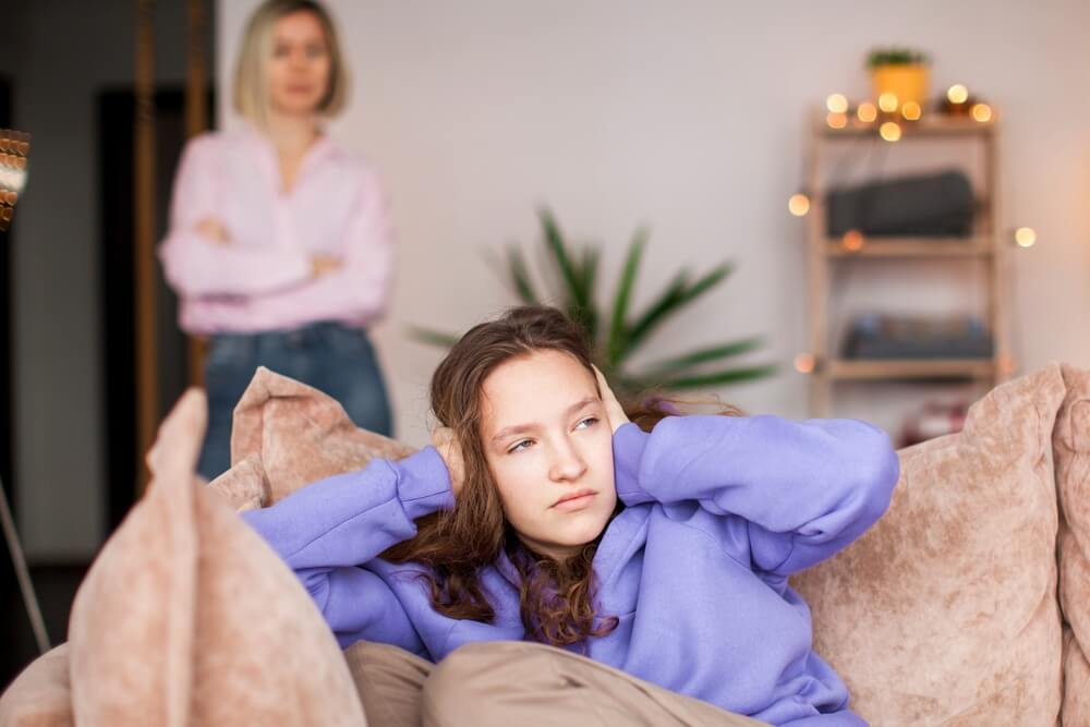 A girl sitting on the couch with both of her hands covering her ears and a women behind her with her arms crossed looking at the girl 