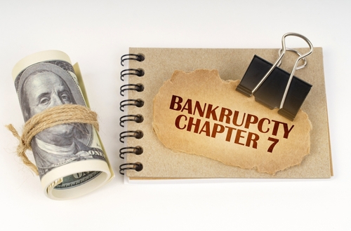A rolled hundred-dollar bill sits beside a notebook featuring a torn paper marked "Bankruptcy Chapter 7," secured with a binder clip—a subtle hint at the looming question: what documents do I need to file for bankruptcies?.