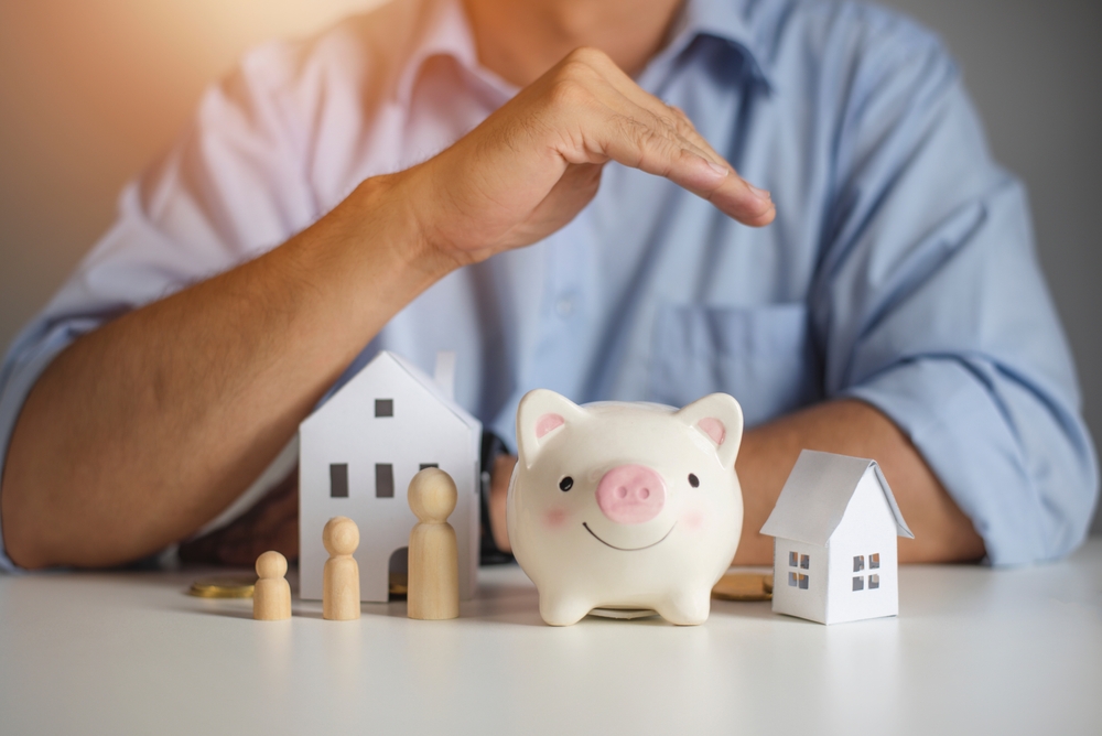 A person’s hand hovers over a piggy bank, with wooden family figures and two small house models on a table
