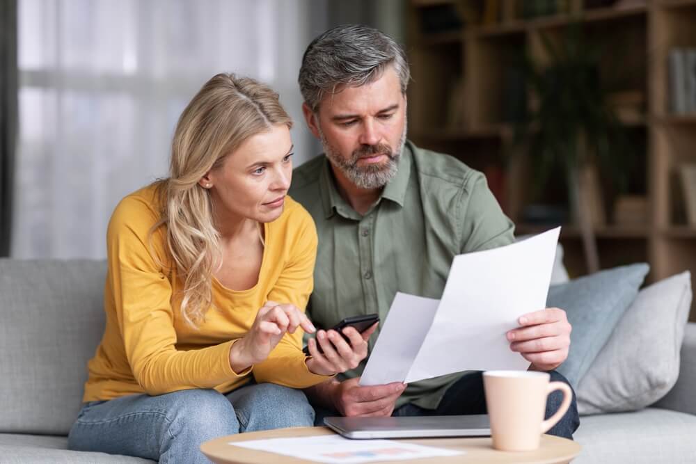 A man and woman sitting on the couch looking at papers and and the woman is holding a calculator 